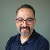 Smiling man with glasses, beard, and mustache wearing a dark polo shirt against a gray background.